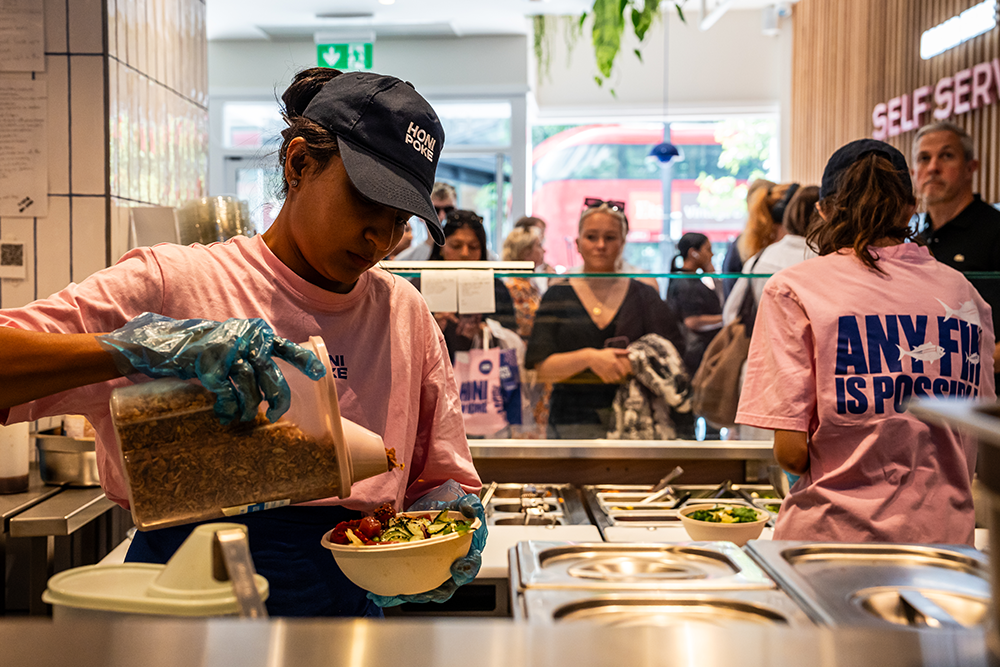 Behind the counter at a poke shop, a kitchen worker creates a poke bowl while customers wait